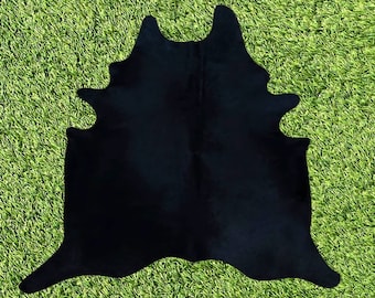 May include: A black cowhide rug with a natural, irregular shape. The rug is laid out on a green grassy surface.