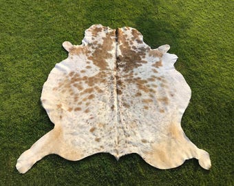 May include: A natural cowhide rug featuring an irregular, organic shape with a white base color and scattered light brown and tan patches. The hide displays a central seam running vertically, with natural edges and a smooth, flat texture.
