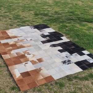 Może przedstawiać: Patchworkowa skóra krowia w postaci dywanu z kwadratami w kolorze brązowym, czarnym, białym i szarym. Dywan leży na zielonej trawie.