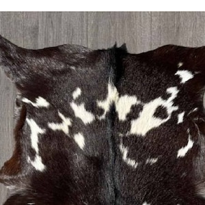 Puede incluir: Una alfombra de piel de vaca negra y blanca con un patr&oacute;n natural moteado. La piel es suave y flexible, con un color marr&oacute;n oscuro intenso.