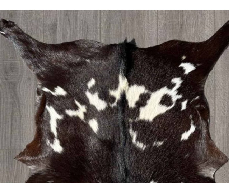 Puede incluir: Una alfombra de piel de vaca negra y blanca con un patr&oacute;n natural moteado. La piel es suave y flexible, con un color marr&oacute;n oscuro intenso.