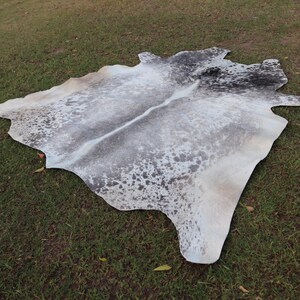 May include: A white cowhide rug with black and gray markings. The rug is laid out on a green grassy surface.