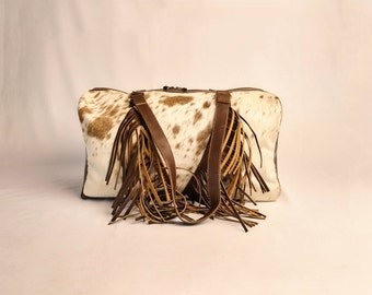 Könnte beinhalten: Eine braune und weiße Kuhfell-Reisetasche mit Fransenbesatz und braunen Lederriemen.