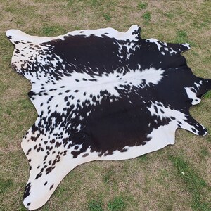 May include: Black and white cowhide rug with a spotted pattern. The rug is laid out on a green grassy surface.
