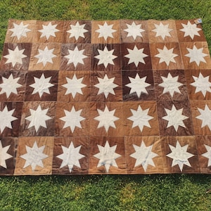 May include: A brown and white cowhide rug with a star pattern. The rug is made up of individual squares of cowhide, each with a white star in the centre. The rug is perfect for adding a touch of rustic charm to any room.