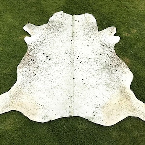 May include: A cowhide rug with a white base and speckled black and gray markings. The rug is irregularly shaped, with a natural edge, and is displayed on a green surface.