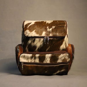 May include: A brown and white cowhide backpack with a flap closure and multiple zipped compartments. The backpack has brown leather accents and silver-toned zips. The bag is designed for carrying items.