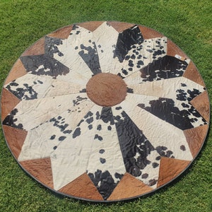 Op de afbeelding: Een rond, bruin, zwart en wit koeienhuidkleed met een stervormig patroon. Het kleed is gemaakt van meerdere stukken koeienhuid die aan elkaar zijn genaaid.