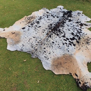 May include: A cowhide rug with black and white spots and a tan patch. The rug is laid out on a green grassy surface.