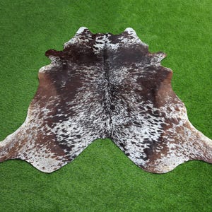 May include: A brown and white cowhide rug with a spotted pattern. The rug is laid out on a green grassy surface.