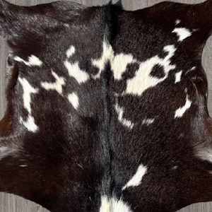 Puede incluir: Una alfombra de piel de vaca negra y blanca con una textura natural y suave. La piel tiene un patr&oacute;n &uacute;nico de manchas blancas sobre un fondo negro.