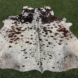 May include: A white cowhide rug with brown and black spots. The rug is laid out on a green grassy surface.