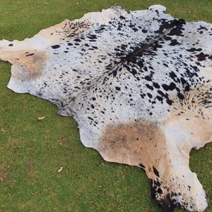 May include: A cowhide rug with a white, black, and brown spotted pattern. The rug is laid out on a green grassy surface.