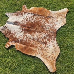 Op de afbeelding: Een bruin en wit koeienhuidkleed met een gevlekt patroon. Het kleed ligt op een groene grasmat.