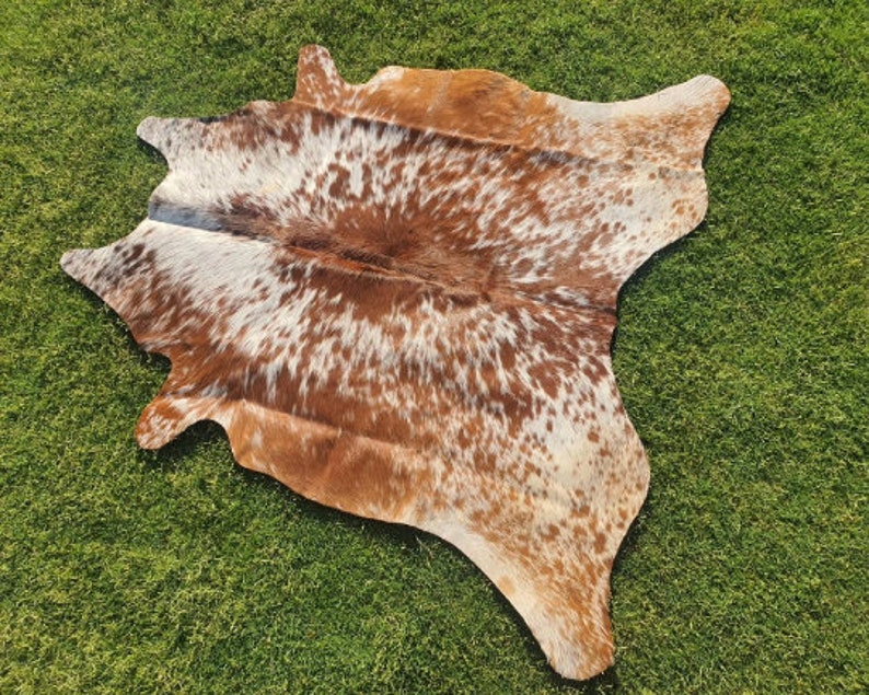 Op de afbeelding: Een bruin en wit koeienhuidkleed met een gevlekt patroon. Het kleed ligt op een groene grasmat.