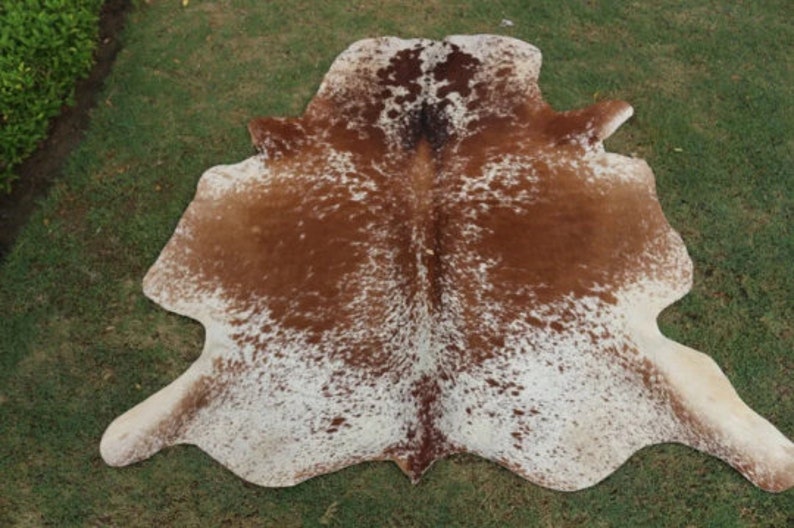May include: A brown and white cowhide rug with a spotted pattern. The rug is laid out on a green grassy surface.