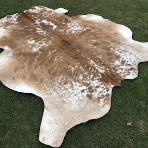 Pode incluir: Um tapete de pele de vaca marrom e branco com um padr&atilde;o natural manchado. O tapete est&aacute; colocado sobre uma superf&iacute;cie de grama verde.