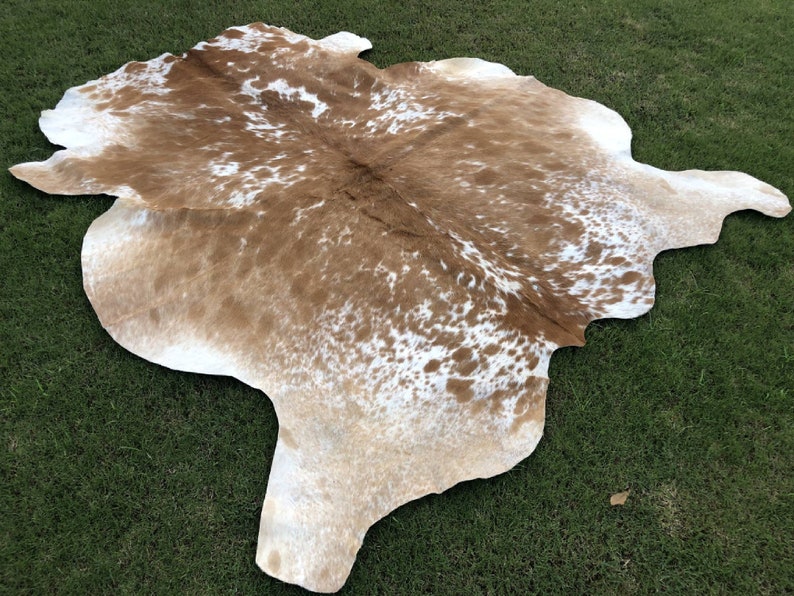 Pode incluir: Um tapete de pele de vaca marrom e branco com um padr&atilde;o natural manchado. O tapete est&aacute; colocado sobre uma superf&iacute;cie de grama verde.