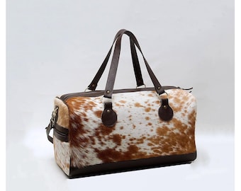 Könnte beinhalten: Eine braune Ledertasche zum Reisen mit einem weißen und braunen Kuhfellmuster. Die Tasche hat zwei obere Griffe und einen Reißverschluss.