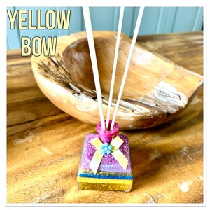 May include: A decorative reed diffuser with a pink and purple glittery bottle, a yellow bow, and a blue flower. The bottle has white reeds and the text "YELLOW BOW" in a yellow box. The diffuser sits on a wooden surface.