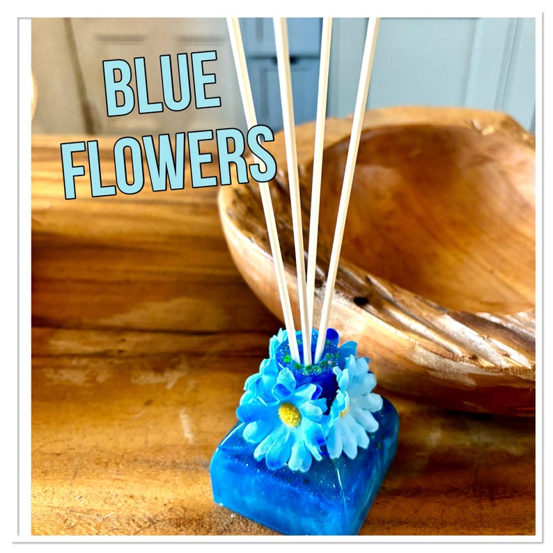 May include: A blue floral diffuser with white reeds. The base is a square of blue resin, decorated with blue and white artificial flowers. The text "BLUE FLOWERS" is displayed in a light blue font.