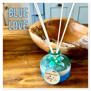 May include: A decorative diffuser bottle with a blue, white, and green design, featuring reed sticks. The bottle has a tag that says "handmade with Love". The words "BLUE LOVE" are in the background.