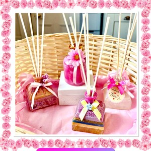 May include: Several decorative reed diffusers in shades of pink and purple. Each diffuser is decorated with ribbons, flowers, and other embellishments. The diffusers are arranged on a pink surface, with a woven basket in the background.