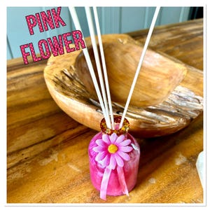 May include: A pink flower-shaped diffuser bottle with white reeds. The bottle has a pink flower and ribbon. The text "PINK FLOWER" is displayed in pink letters with white polka dots. The diffuser sits on a wooden surface.