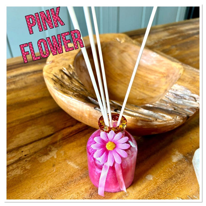 May include: A pink flower-shaped diffuser bottle with white reeds. The bottle has a pink flower and ribbon. The text "PINK FLOWER" is displayed in pink letters with white polka dots. The diffuser sits on a wooden surface.
