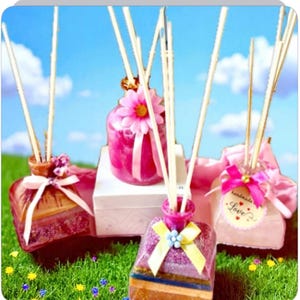 May include: A collection of decorative reed diffusers in shades of pink and purple, with wooden reeds. Each diffuser is decorated with ribbons, flowers, and tags. The background shows a blue sky and green grass.