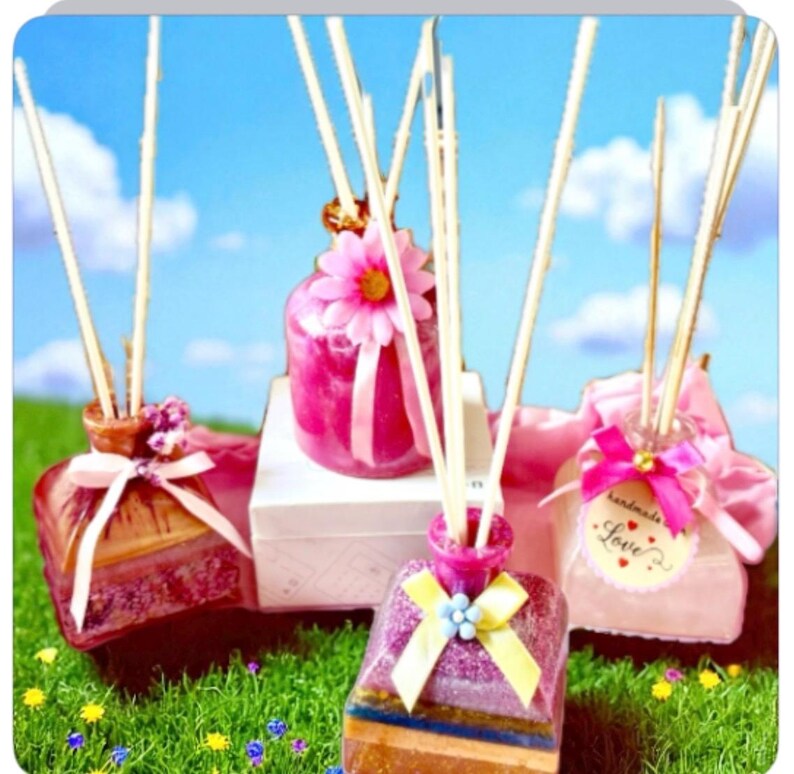 May include: A collection of decorative reed diffusers in shades of pink and purple, with wooden reeds. Each diffuser is decorated with ribbons, flowers, and tags. The background shows a blue sky and green grass.