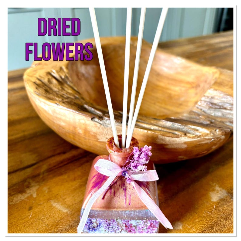 May include: A decorative reed diffuser with the text "DRIED FLOWERS". The diffuser has a pink ribbon and dried flowers. White reeds are inserted into the bottle. A wooden bowl is in the background.