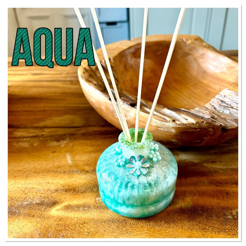 May include: An aqua-coloured diffuser bottle with a floral design and wooden reeds. The word "AQUA" is displayed in large, teal letters. The bottle sits on a wooden surface, with a wooden bowl in the background.