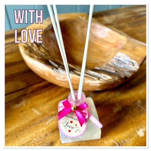 May include: A handmade aromatherapy diffuser with reed sticks. The small glass bottle is decorated with a pink ribbon and a tag that says "handmade" and "Love". The words "WITH LOVE" are in pink text in the background. The diffuser sits on a wooden surface.