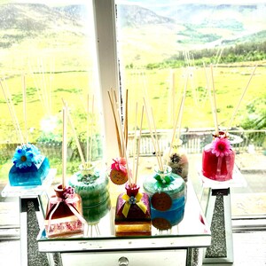 May include: A selection of colourful reed diffusers with decorative tops and wooden sticks. The diffusers are displayed on a mirrored table and individual stands, with a scenic outdoor view in the background.
