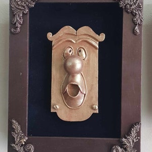 Peut inclure: Une œuvre d'art murale encadrée fantaisiste représentant un visage stylisé de couleur or avec une moustache et une bouche ouverte. L'œuvre est placée sur un fond noir dans un cadre marron avec des détails d'angle ornés.