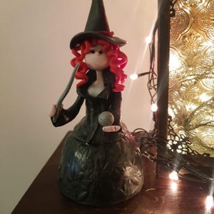 Puede incluir: Figura de bruja artesanal con pelo rizado rojo, sombrero negro y vestido negro. La bruja sostiene una varita y una bola de cristal. La figura es un objeto decorativo, posiblemente para Halloween o un tema de fantasía.
