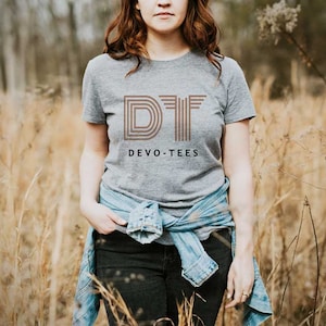 Op de afbeelding: Een vrouw draagt een grijs T-shirt met een bruine afbeelding waarop "DIT DEVO-TEES" staat. Ze staat in een veld met hoog gras.