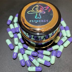 May include: A container with a gold rim and the word "PSYNERGY" on the lid, featuring a mushroom design. Numerous purple and light green capsules are scattered around the container, creating a vibrant arrangement.