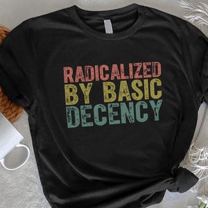 May include: Black t-shirt with the text "RADICALIZED BY BASIC DECENCY" in a retro font. The text is in red, yellow, and teal. The shirt is on a white surface with a woven basket, a white mug, and flowers.