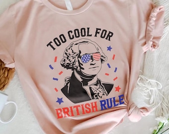 Può includere: T-shirt rosa chiaro con una grafica di George Washington con occhiali da sole con il design della bandiera americana. Il testo recita "TOO COOL FOR BRITISH RULE" in rosso, bianco e blu.