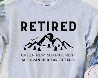 May include: A pale grey sweatshirt featuring the word "RETIRED" in black, above a black mountain and tree graphic. The text "UNDER NEW MANAGEMENT SEE GRANDKID FOR DETAILS" is printed below the graphic. The sweatshirt is paired with blue jeans and white trainers.
