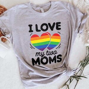 May include: A light gray t-shirt with the text "I LOVE my two MOMS" in black. The design features two rainbow-colored hearts. The shirt is styled with a knot at the bottom and is next to a white mug and flowers.