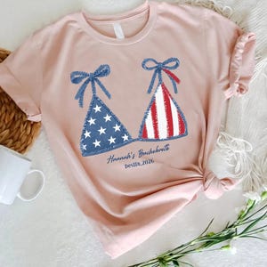 May include: A light pink t-shirt featuring a graphic of two bikini tops with blue bows, one with white stars on a blue background and the other with red and white stripes. The text "Hannah's Bachelorette Destin 2026" is printed below the graphic.