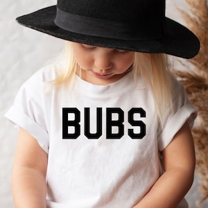 May include: A young child wearing a white t-shirt with the word "BUBS" in bold black letters. The child is also wearing a black hat and brown pants. The child is holding a small bundle of dried flowers.