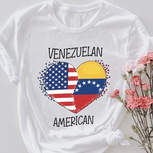 May include: White t-shirt featuring a heart-shaped design combining the American and Venezuelan flags. The words "VENEZUELAN" and "AMERICAN" are printed above and below the heart, respectively. A bouquet of pink flowers is to the right.