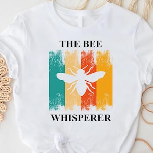 May include: White t-shirt with the text "THE BEE WHISPERER" above and below a white bee silhouette. The bee is set against a background of vertical stripes in teal, yellow, orange, and red. The shirt is knotted at the waist.