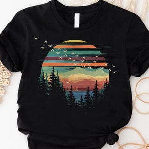 May include: Black t-shirt featuring a colorful graphic of a sunset over a mountain range with pine trees. The design includes a vibrant sky with birds in flight, creating a nature-inspired scene.