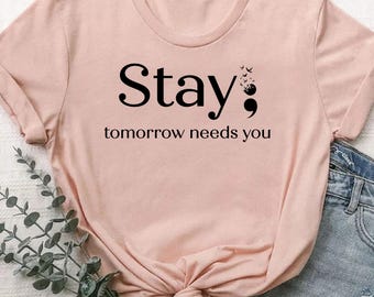 May include: A heather peach t-shirt with the words "Stay; tomorrow needs you" printed in black. The shirt has short sleeves and a crew neck. The shirt is paired with denim shorts.
