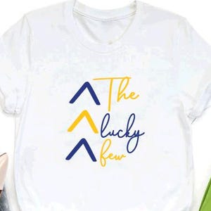 May include: White t-shirt with a blue and yellow graphic that says "The lucky few" with three blue and yellow arrows pointing upwards.
